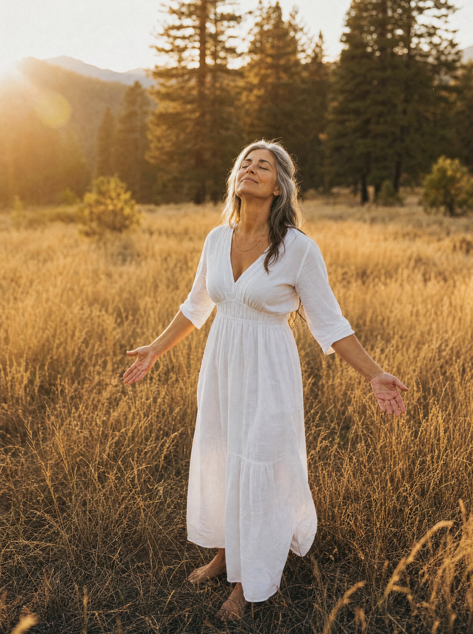 A silver-haired woman standing in a golden meadow, face lifted to the light, arms loose and open