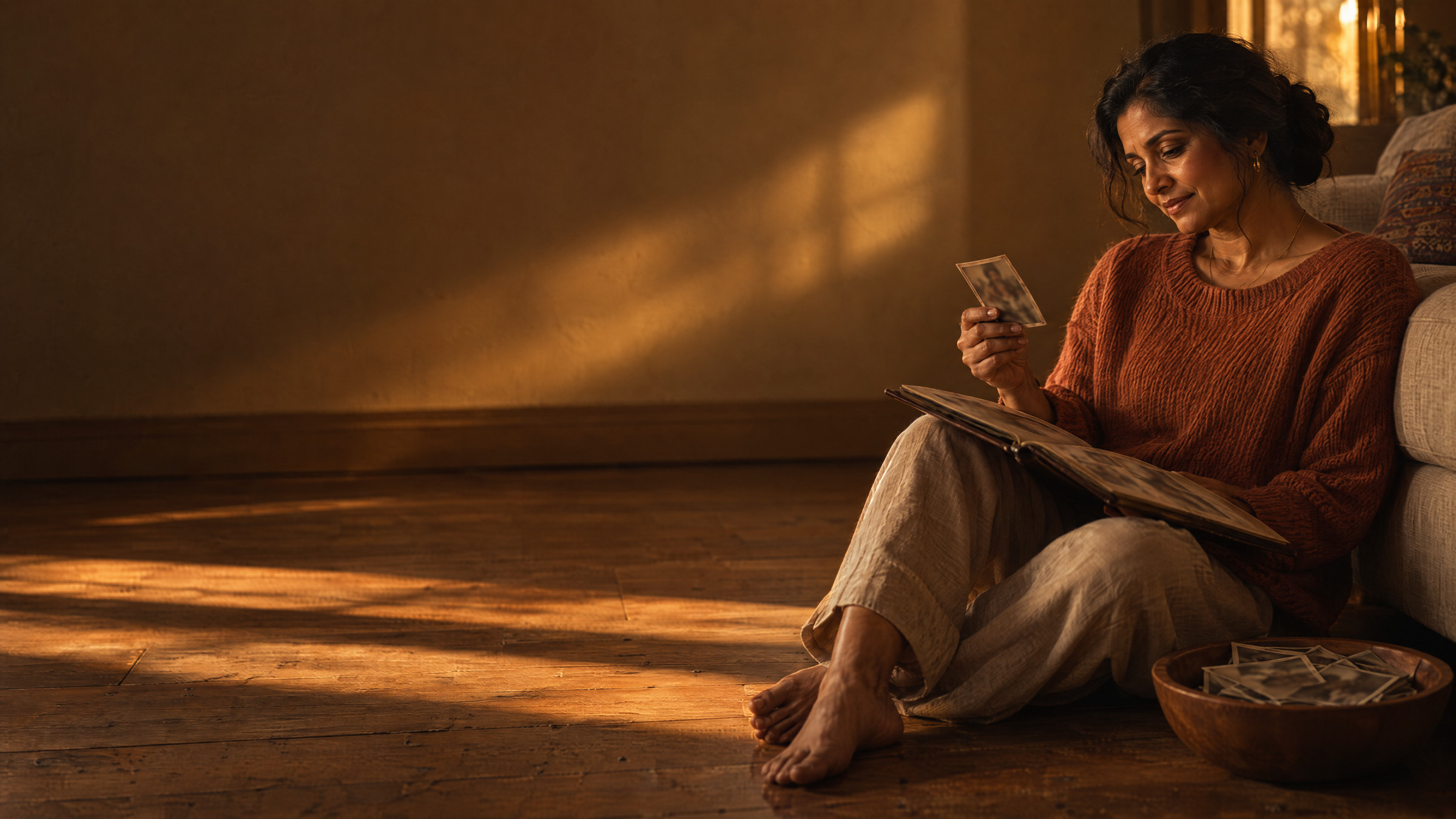 A woman sitting on a sunlit wooden floor with an old photo album open in her lap, studying one photograph