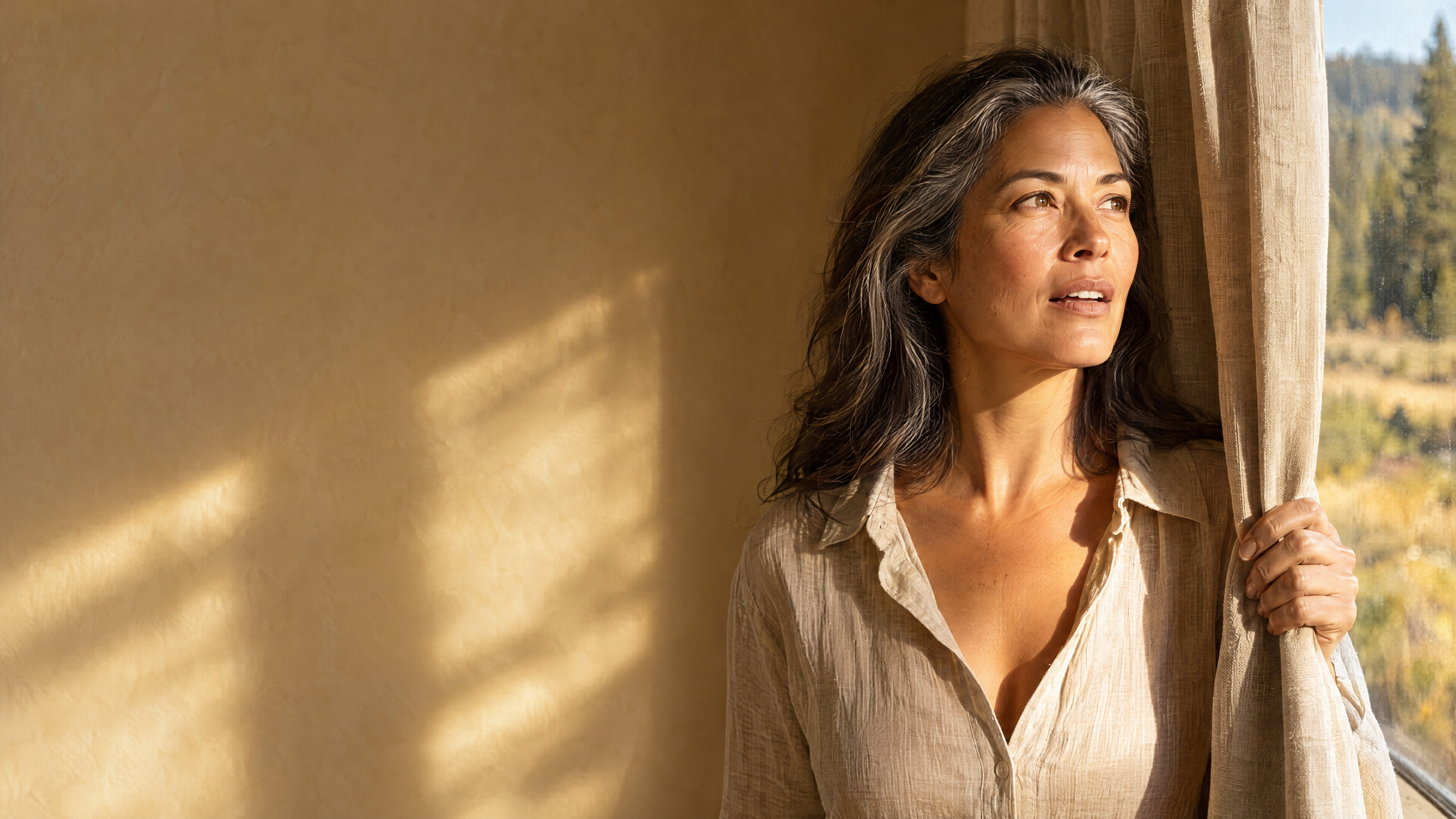 A woman drawing a linen curtain aside as morning sun floods across her face