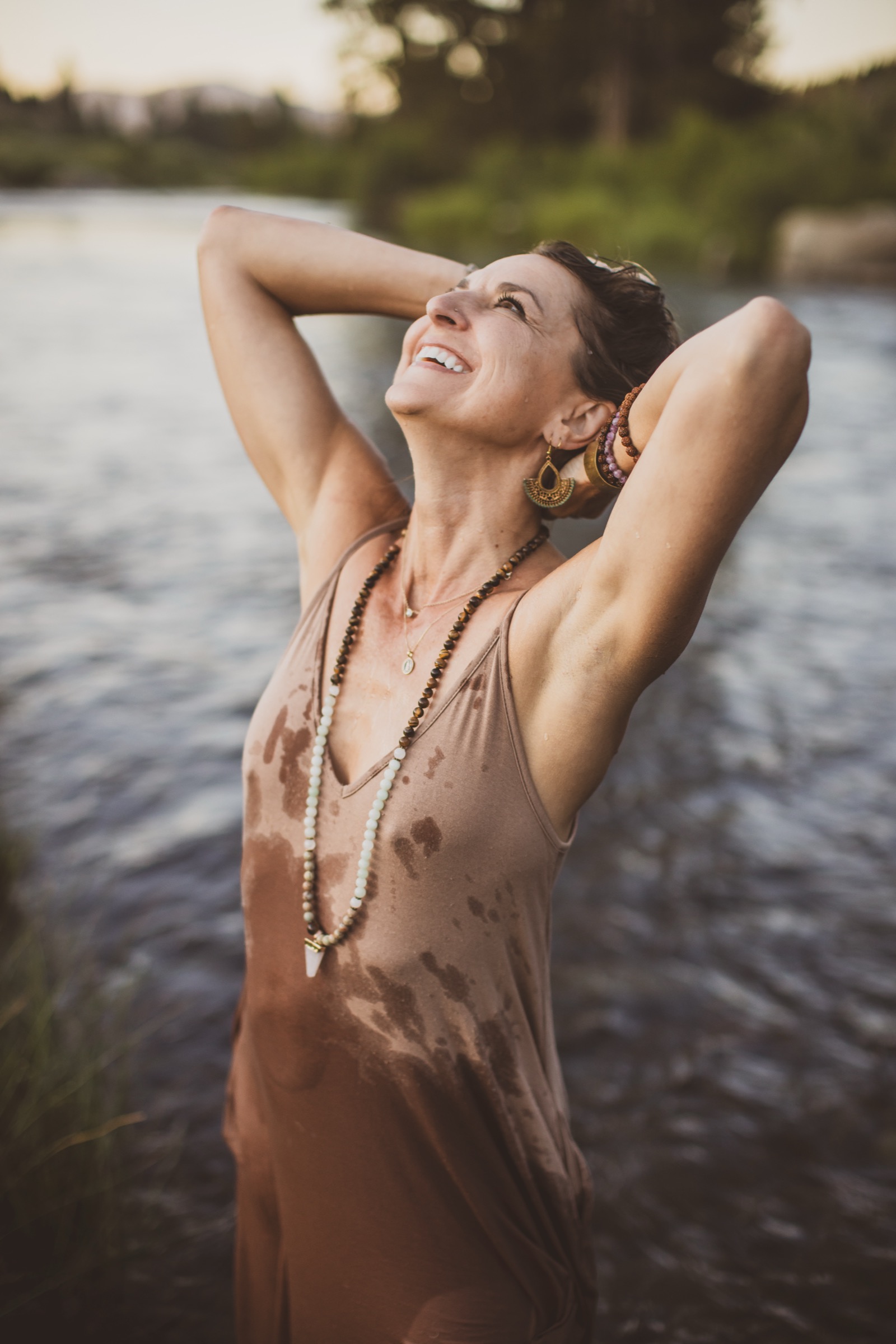 Kaila standing in the river at sunset, arms raised behind her head, laughing